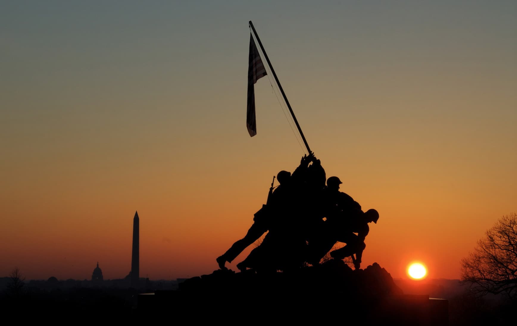 The Marine Corps War Memorial in silhouette at sunset, the Washington Monument and the Capitol dome on the horizon behind it.